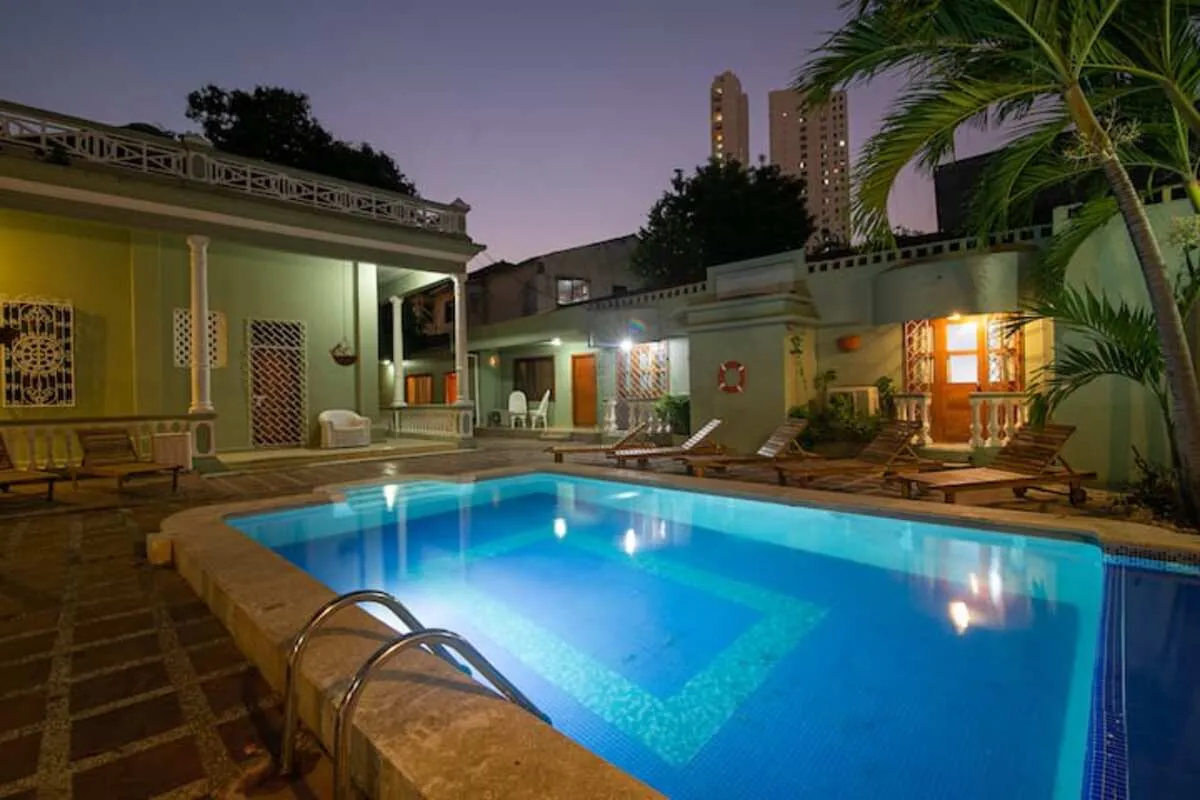 Piscina azul iluminada junto a palmeras y fachada amarilla clasica